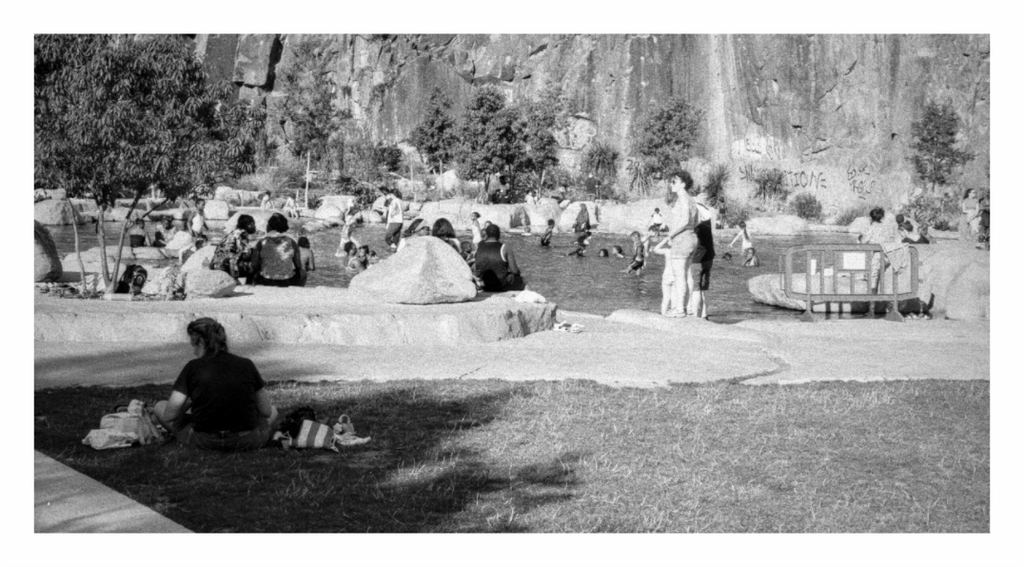 People sitting on grass and white rock ledges beside the quarry pool at the Jardin Extraordinaire in Nantes, cliff face behind, black and white