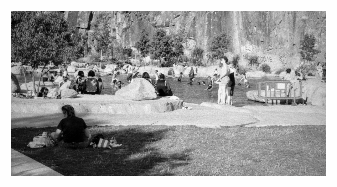 People sitting on grass and white rock ledges beside the quarry pool at the Jardin Extraordinaire in Nantes, cliff face behind, black and white