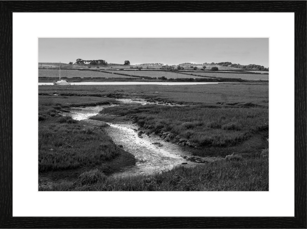 A tidal creek winding through salt marsh grass in the Aln estuary at Alnmouth, a small white boat on the water beyond, black and white
