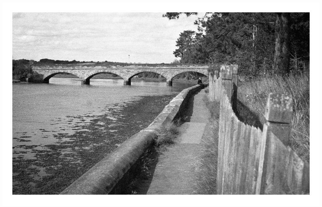 The three-arch stone bridge over the River Aln at Alnmouth, a figure sitting on the parapet, black and white