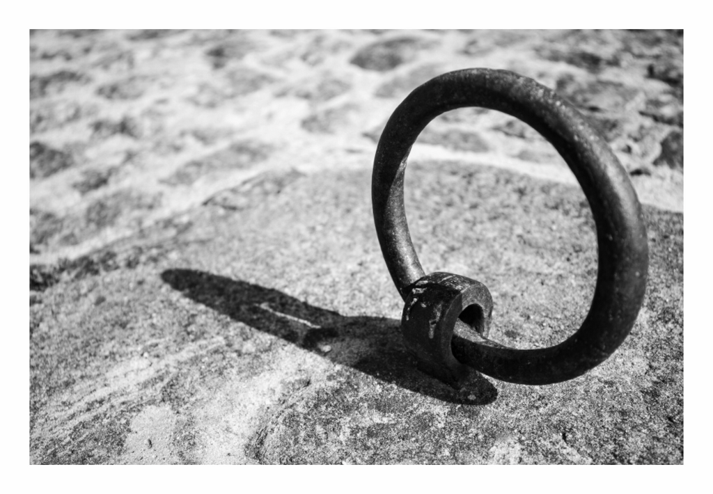 A hand-forged iron mooring ring bolted into the stone quay at Montjean-sur-Loire, casting a hard shadow across the granite, black and white