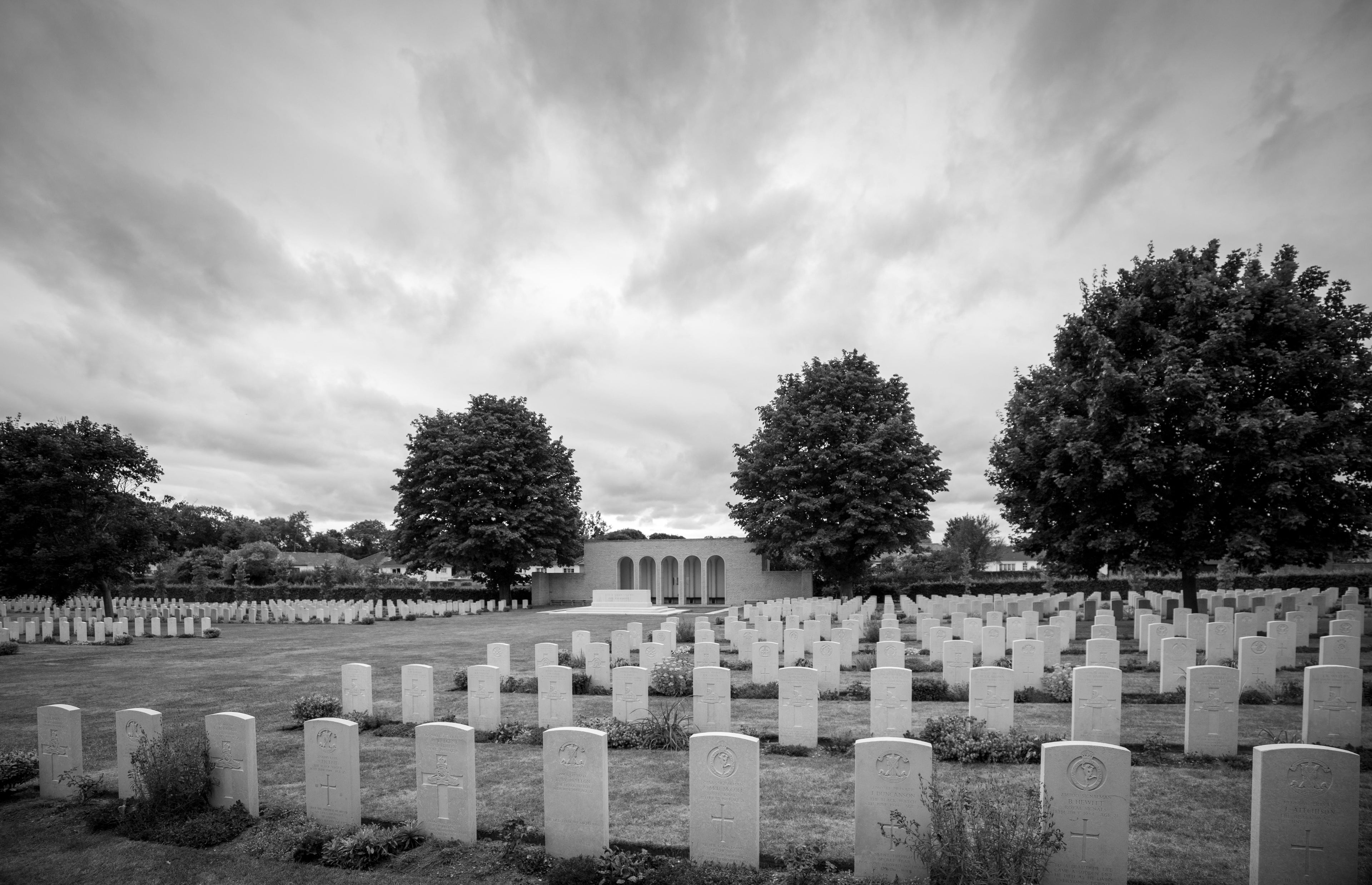 Rows of Commonwealth headstones running away towards the cemetery shelter and trees under heavy cloud, Ranville War Cemetery, Normandy