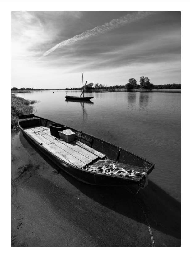 A flat-bottomed wooden Loire boat drawn up on sand, a masted boat at anchor beyond it and streaked cloud above, black and white